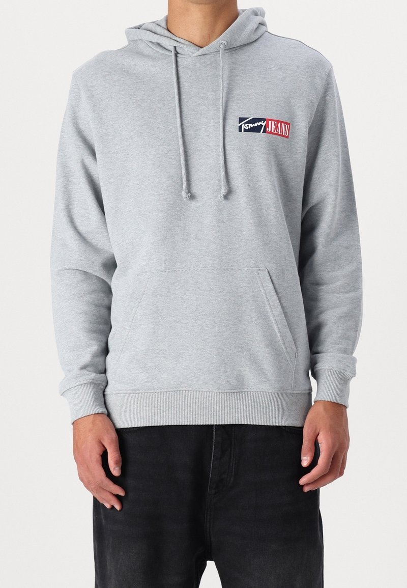 Homme portant un sweat à capuche gris clair avec poche avant et logo Tommy Jeans sur la poitrine, associé à un pantalon noir, debout devant un fond uni.