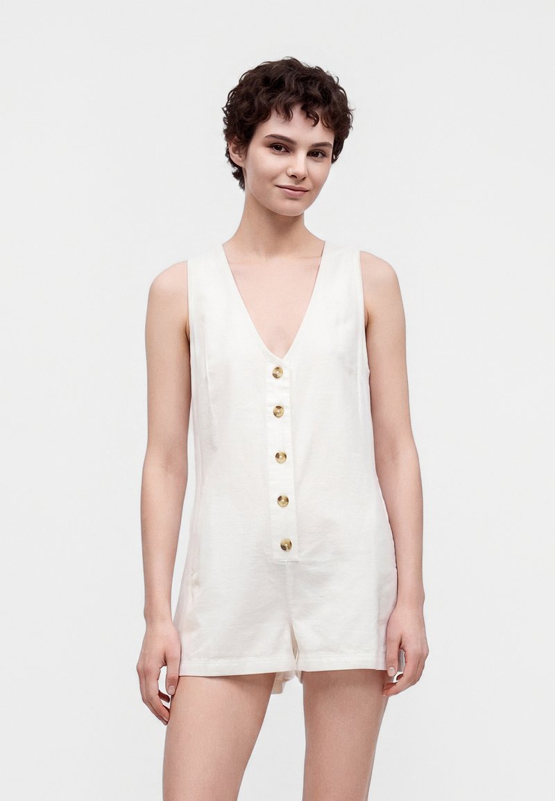 Femme aux cheveux courts bouclés portant une combinaison blanche sans manches boutonnée, debout devant un fond clair uni, souriant légèrement.
