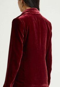 Femme portant un blazer en velours rouge profond, vue de dos, avec des cheveux foncés attachés et une boucle d'oreille en or pendante visible à l'oreille gauche.