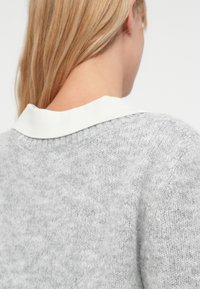 Blondais cilvēks skatīts no aizmugures, ģērbies gaiši pelēkā adītā džemperī ar baltu apkakli uz vienkārša fona.