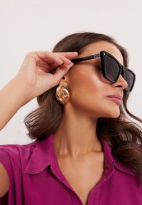 Czarne prostokątne okulary przeciwsłoneczne z ciemnymi soczewkami; duże złote abstrakcyjne kolczyki; nosi fioletową koszulę z kołnierzykiem; gładka struktura włosów.