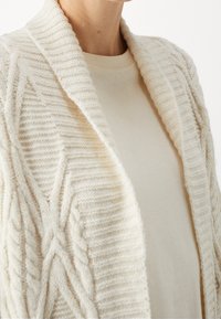 Detailní záběr osoby v krémovém kardiganu s copánkovým vzorem, který má na sobě odpovídající tričko s kulatým výstřihem, na pozadí s neutrální barvou.