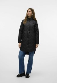 Zwarte gewatteerde lange jas met hoge kraag, knoopsluiting en zijzakken, gecombineerd met blauwe recht gesneden jeans en zwarte schoenen.
