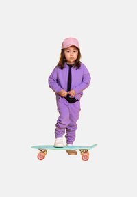 Fioletowy komplet dresowy z czarną bluzką. Na głowie różowa czapka, stoi na turkusowej deskorolce z różowymi kółkami.