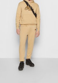 Beige Jogginghose und Kapuzenpullover mit schwarzem Nike-Logo. Der Pullover verfügt über eine Fronttasche; die Hose hat das passende Logo. Schwarze Sportschuhe.