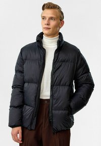 Doudoune noire avec un col haut, dotée d'une fermeture éclair et d'une texture matelassée. Associée à un col roulé blanc en dessous.