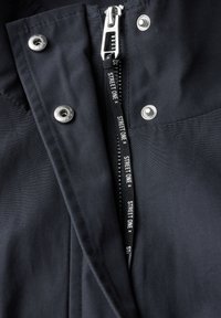 Zwarte jas met een zilveren ritssluiting, drukknoopsluiting en een stoffen label met "STREET ONE". Gladde textuur en taillerend ontwerp.