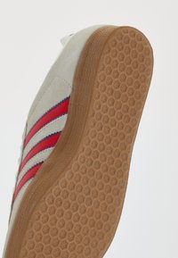 Adidași cu un superior din suede bej, având trei dungi roșii și accente albastre. Talpă din cauciuc de tip gumă cu un design de profiluri pentru aderență.