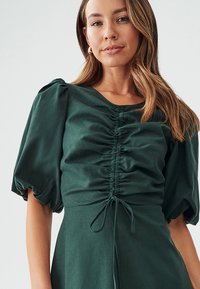 Dark green dress with puffed sleeves, ruched front, and a drawstring waist. Made of soft fabric with a smooth texture.