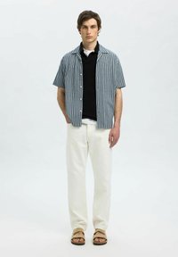 Mėlynos ir baltos dryžuotos trumpomis rankovėmis marškiniai, uždėti ant juodo megzto viršaus, derinami su baltais kelnėmis ir rudais sandalais.