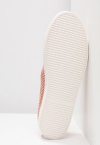 Semelle en caoutchouc blanche avec un motif ondulé, présentée debout contre un mur blanc, avec une partie de la tige de chaussure rose perforée visible.