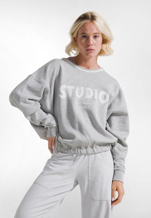 Młoda kobieta z krótkimi blond włosami nosi luźną szarą bluzę z tekstem "STUDIO DEHA" oraz jasne spodnie na białym tle.