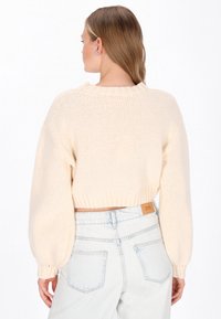 Krótki sweter w kolorze kremowym z prążkowanym wykończeniem i szerokimi rękawami, w zestawieniu z jasnoniebieskimi dżinsami z skórzaną metką i tylnymi kieszeniami.