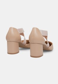 Beige heeled sandals with a rounded toe, smooth leather upper, and thick block heel. Features elastic straps crossing at the ankle.