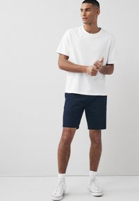 Camiseta de algodón blanca de manga corta combinada con pantalones cortos azul marino. El modelo se presenta con confianza, vistiendo zapatillas altas blancas y calcetines blancos.