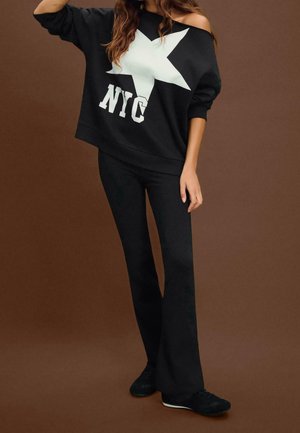 Nainen mustassa olkapäät paljastavassa yläosassa, jossa valkoinen tähti ja "NYC"-teksti, yhdistettynä mustiin leveneviiin housuihin ja mustiin lenkkareihin, seisomassa ruskeaa taustaa vasten.