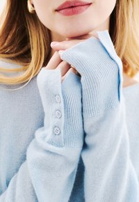 Lichtblauwe trui met geribde mouwen, voorzien van drie kleine knopen op de manchetten. Zachte textuur, losse pasvorm en off-shoulder ontwerp.