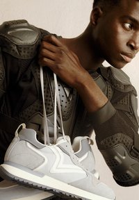 Baskets de sport gris clair en matériau suédé, avec des accents blancs et une semelle grise texturée. Présentées avec des équipements de protection en vue rapprochée.