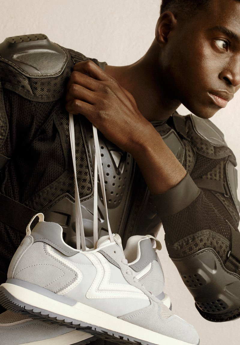 Baskets de sport gris clair en matériau suédé, avec des accents blancs et une semelle grise texturée. Présentées avec des équipements de protection en vue rapprochée.