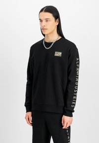 Fekete kapucnis pulóver, amelyen álcázott logófogdosó található a mellkason, és aranyszínű "ALPHA INDUSTRIES" felirat található a bal ujjon. Simán tapintású.