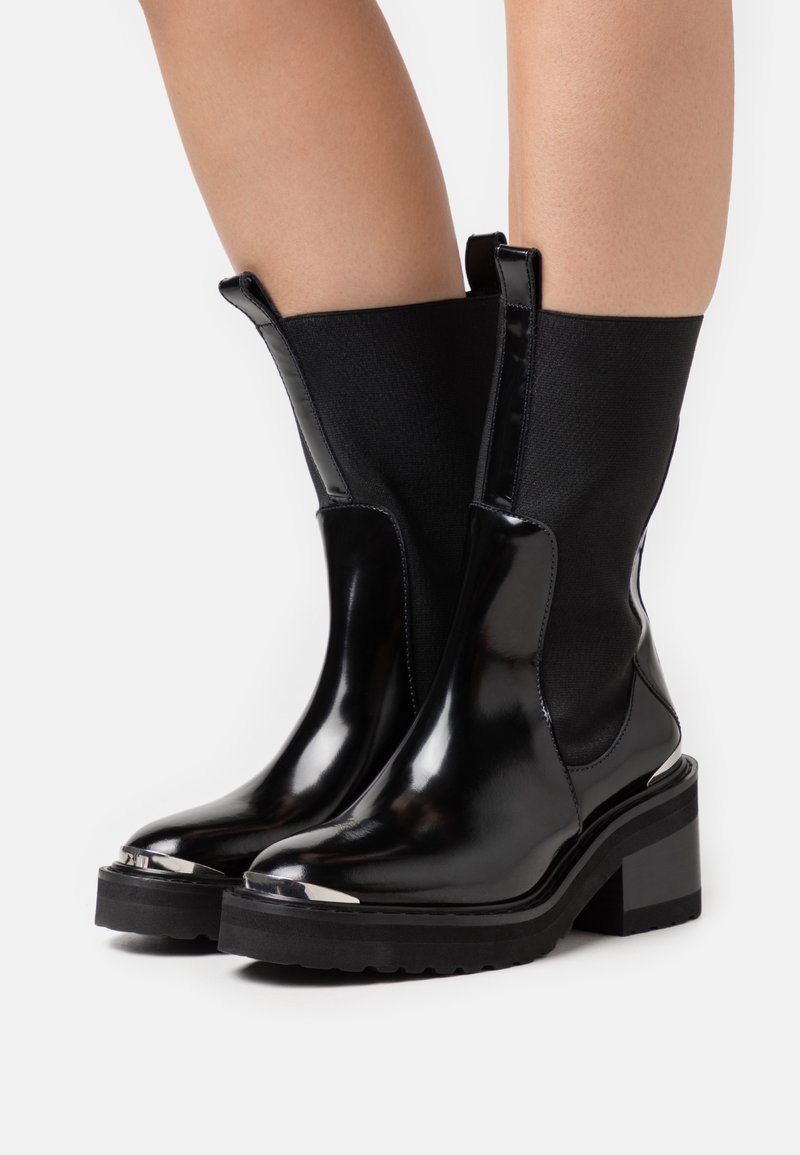 Bottines noires brillantes à talons épais, avec panneaux latéraux élastiques et languettes d’aide à l’enfilage, portées sur jambes nues sur un fond blanc uni.