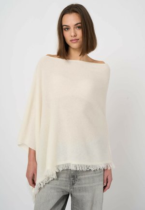 Jonge vrouw met recht, bruin haar die een crèmekleurige off-shoulder poncho met franjes en lichtgrijze spijkerbroek draagt, tegen een witte achtergrond.