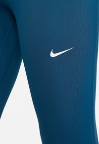 Teal träningsleggings tillverkade av elastisk tyg med platta sömmar och en liten vit Nike-logotyp på vänster sida.