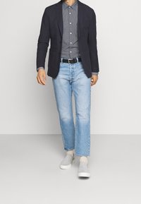 Marinblå blazer, ljusblå denimjeans, grå skjorta med knappar, svart bälte och grå slip-on skor. Avslappnad, modern passform med raka jeansben.