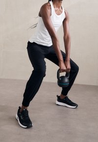 Svarta träningsbyxor och en vit linne tröja bärs medan en svart kettlebell hålls. Svarta sneakers med vita sulor fulländar outfiten.