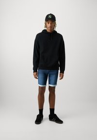 Hanorac negru cu buzunar tip cangur, pantaloni scurți din denim albastru cu margini răsucite, adidași negri și o șapcă neagră cu detalii verzi ale logo-ului.