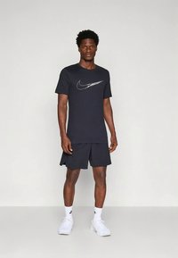 Mees, kes kannab musta Nike t-särki, musti lühikesi pükse, valgeid Nike sokke ja valgeid tossusid, seisab ühevärvilise heleda tausta ees.