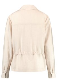 Jasnobeżowa bluzka, lekka tkanina, luźny krój z marszczonym detalem w talii, długie rękawy i klasyczny kołnierzyk. Prosta, gładka faktura.