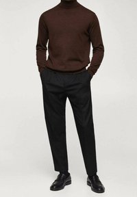 Maglione a collo alto marrone abbinato a pantaloni neri. Il maglione ha un design aderente, mentre i pantaloni presentano pince e gamba dritta. Scarpe nere completano il look.