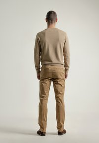 Hombre de pie de espaldas, vistiendo un suéter beige, pantalones color marrón claro y zapatos marrones, frente a un fondo claro y liso.