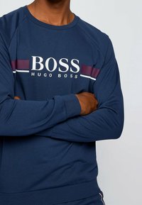 Sudadera azul marino de manga larga, con un logo blanco de "BOSS" y franjas en burdeos y blanco en el pecho. Textura suave, ajuste casual.