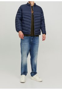 Uomo che indossa una giacca sfoderata blu navy, una maglietta verde oliva, jeans blu sbiaditi e sneaker bianche, in piedi su uno sfondo grigio uniforme.