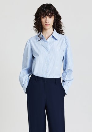 Donna con capelli ricci e scuri, che indossa una blusa a strisce blu chiaro con piccole ricami floreali bianchi e pantaloni navy su misura, con le mani nelle tasche.