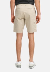 Beige cargo šortky majú bočné a zadné vrecká, sú vyrobené z odolnej látky s voľným strihom a lemom po kolená. Skombinované so šedými topánkami.