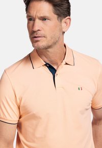 Polo shirt orange clair avec un col et des bordures bleu nuit. Présente un petit logo brodé en vert, blanc et rouge. Manches courtes, coupe classique.