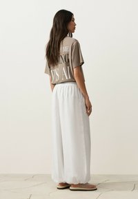 Femme aux longs cheveux foncés portant un t-shirt beige imprimé, un pantalon large blanc et des sandales beiges, debout de profil sur un sol carrelé clair.