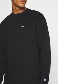 Sudadera negra de cuello redondo hecha de tela suave, con un pequeño logo en el pecho y un detalle de bordado en contraste en la manga.