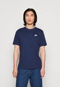 Jūras zila kokvilnas t-krekls ar apaļu kaklu un īsām piedurknēm; tam ir neliels balts Nike logotips kreisajā krūšu daļā.