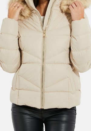 Kvinde iført beige quiltet dunjakke med pelsforet hætte, lynlås foran, og sorte skindbukser, der holder hætten med begge hænder.