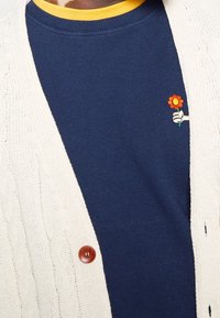 Knit cream cardigan with cable pattern, wooden buttons; layered over a navy blue long-sleeve shirt featuring a small orange flower embroidery.