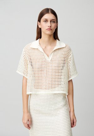 Polo shirt blanc à manches courtes avec un col en V, présentant une texture tissée. Assortie à une jupe dans un motif similaire.