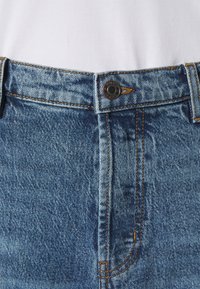 Jean en denim bleu avec une coupe taille haute, détail de bouton en bronze, et coutures apparentes. Le matériau présente une finition classique et texturée.