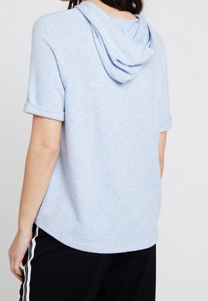 Femme portant un sweat à capuche bleu clair à manches courtes avec des poignets retroussés et un pantalon noir avec des bandes blanches sur les côtés, vue de dos.
