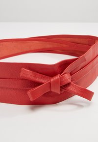 Cintura in pelle rossa con una finitura testurizzata, caratterizzata da un accento annodato e da diverse cinghie sottili per una vestibilità regolabile.