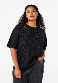 Una donna con i capelli ricci indossa una maglietta nera a maniche corte con piccoli punti bianchi e jeans grigio scuro, con una mano in tasca e un'espressione neutra.
