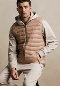 Beżowa kamizelka puchowa z poziomym przeszyciem, kapturem i suwakiem z przodu. Połączona z jasnobeżową bluzą i dopasowanymi spodniami. Zauważalny detal kieszeni.